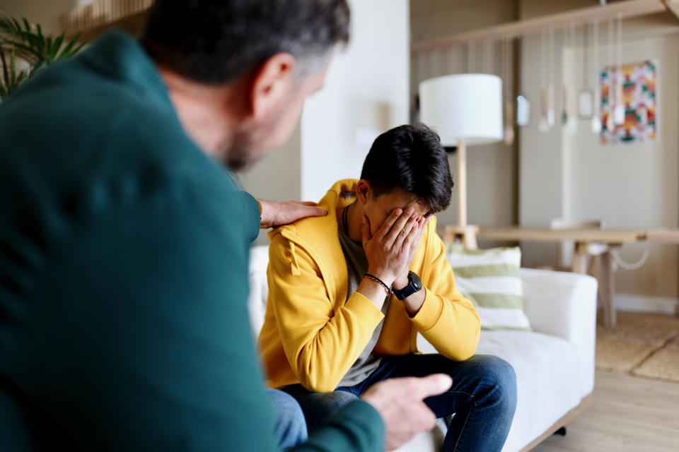 A man in a yellow hoodie on a sofa covers his face as another man comforts him, patting his shoulder